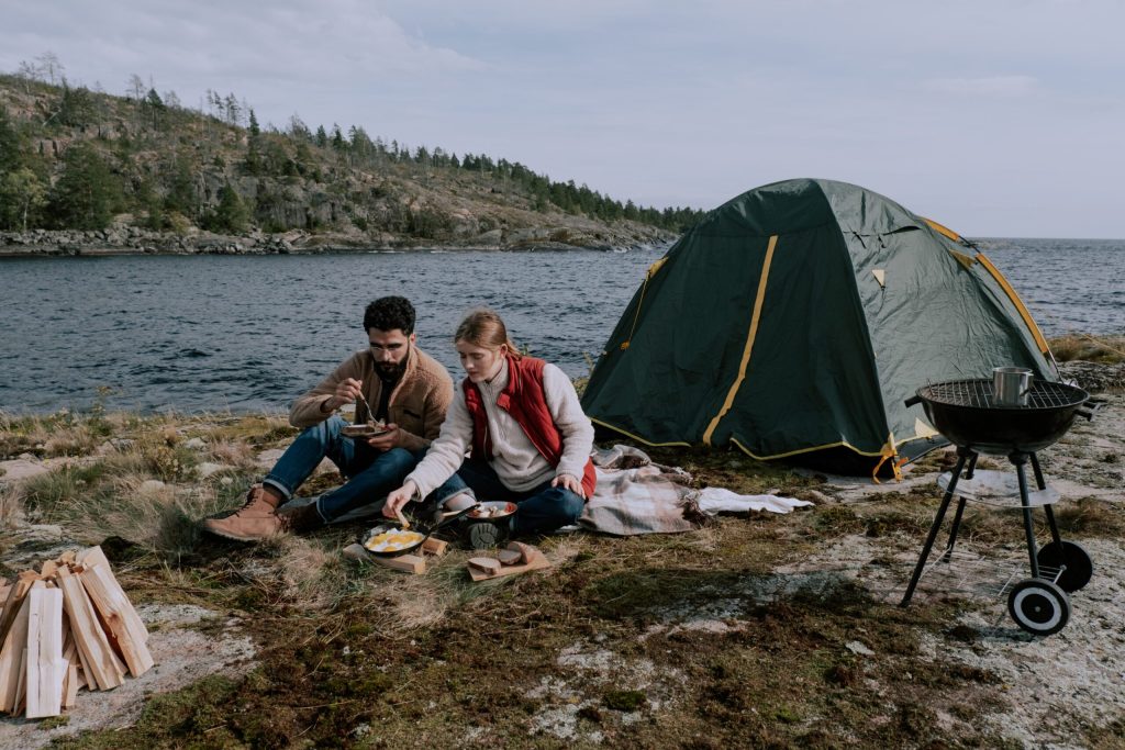 a-couple-sitting-on-picnic-mat-near-a-tent-along-a-river